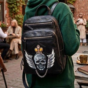 May include: A black leather backpack featuring a graphic print of a skull with white wings and a gold crown on the front pocket. The backpack includes two zippered compartments, a top handle, and a silver-toned chain detail below the skull.