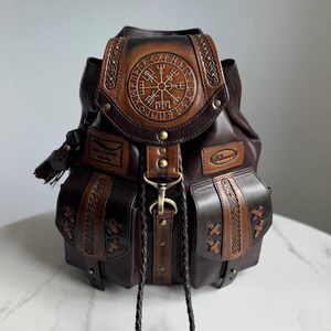 Pode incluir: Uma mochila de couro castanho escuro com um símbolo Vegvisir circular em relevo na aba superior. A bolsa inclui dois bolsos frontais com detalhes em couro trançado, um fecho central de latão e dois longos cordões de couro trançado.