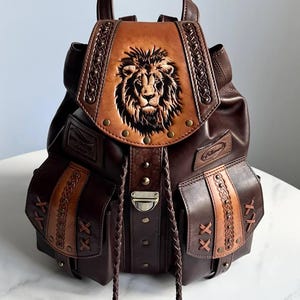 Op de afbeelding: Bruin leren rugzak met een leeuwenkop in reliëf op de flap. De rugzak heeft twee zijzakken, decoratieve stiksels en gevlochten trekkoorden. Hij heeft een handvat aan de bovenkant en verstelbare bandjes.