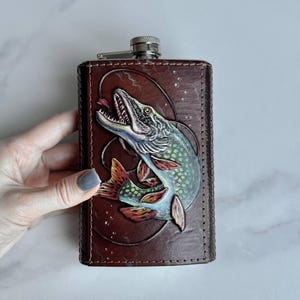 Può includere: Una fiaschetta in pelle marrone con un'immagine in rilievo di un pesce. Il pesce è raffigurato con sfumature di blu, rosso e verde. La fiaschetta ha un tappo argentato ed è di forma rettangolare.