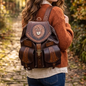 May include: A brown leather backpack featuring a detailed embossed lion head on the top flap, two front pockets with intricate braided trim, brass-colored metal hardware, a central buckle closure, and adjustable shoulder straps for carrying.