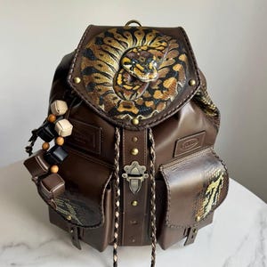 Puede incluir: Una mochila de cuero marrón con una solapa circular que presenta una ilustración detallada de una serpiente. La bolsa incluye dos bolsillos frontales con detalles de piel de serpiente, un cierre central de metal, cordones trenzados y cuentas de madera.