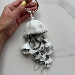 May include: A white leather jellyfish-shaped keychain with a silver ring. The jellyfish has a fringed edge and layered, leaf-like details. A small pearl bead is attached to the top.