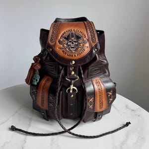 May include: A dark brown leather backpack featuring a tan flap with an embossed skull wearing a pirate hat, crossed swords, and the text "THINK LIKE A CAPTAIN ACT LIKE A PIRATE." The bag includes braided drawstrings, side pockets, and a small skull charm.