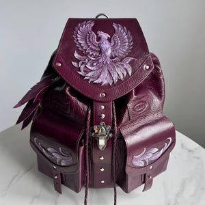 Może przedstawiać: Bordowy skórzany plecak ze szczegółowym fioletowym wzorem feniksa na klapie. Plecak posiada dwie boczne kieszenie z dekoracyjnymi akcentami, srebrne zapięcie i pióro. Torba ma teksturowane wykończenie.