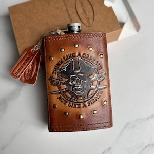 May include: A brown leather hip flask with a pirate design. The flask has a metal skull wearing a captain's hat, crossed swords, and the text "THINK LIKE A CAPTAIN ACT LIKE A PIRATE". It is decorated with brass studs and a small leather tag.