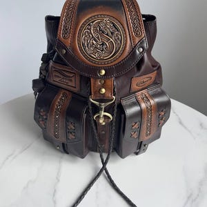 May include: A dark brown leather backpack featuring a circular embossed dragon design on the top flap, braided leather accents, two front pockets with crisscross lacing, brass hardware, and a central metal clasp with two dangling braided leather cords.