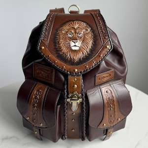 May include: Brown leather backpack with a detailed lion's head design on the flap. The backpack features two front pockets, decorative stitching, and a metal clasp. The bag has a top handle and adjustable straps.