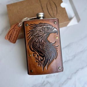 May include: A brown leather hip flask featuring an embossed eagle design. The flask has a silver cap and brass rivets. A small tag with "hand made" is attached. The flask is rectangular and comes with a brown cardboard box.