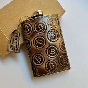 Puede incluir: Petaca de cuero marrón con un diseño de símbolos Bitcoin en relieve. La petaca tiene un tapón plateado y una pequeña etiqueta que dice "hand made". Presentada con una caja de cartón marrón.