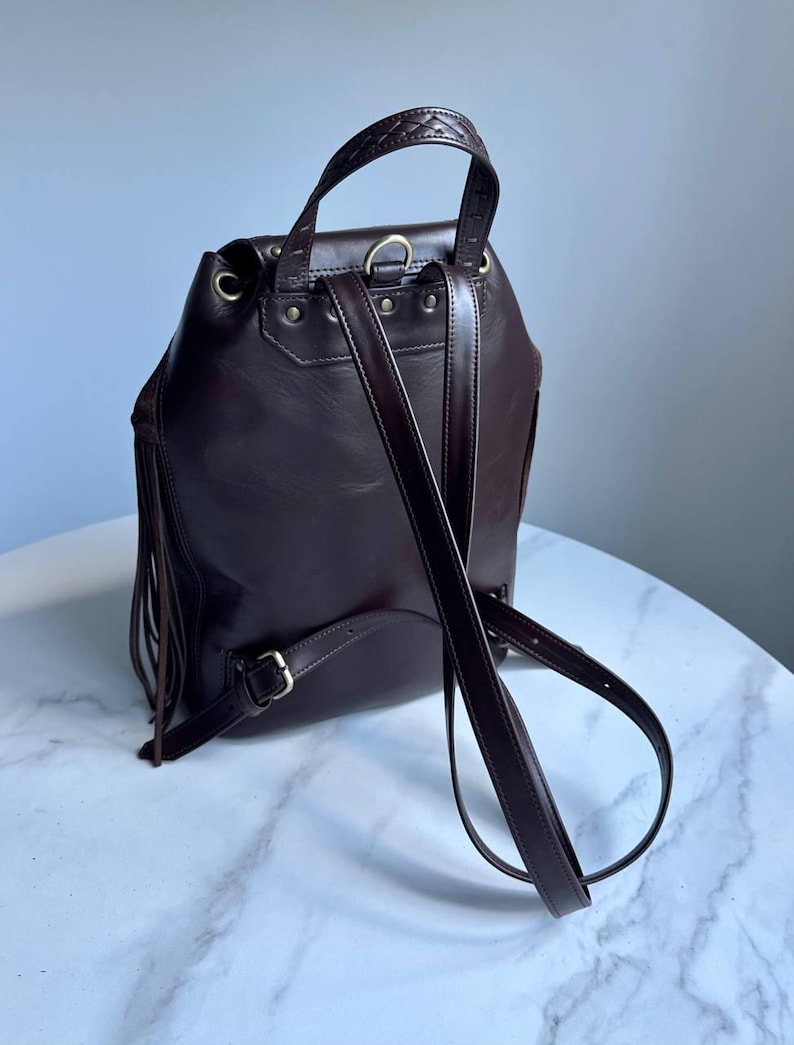 Pu&ograve; includere: Uno zaino in pelle marrone scuro con lunghe frange sui lati, un manico superiore con cuciture intrecciate, spallacci regolabili e minuteria metallica color ottone anticato, inclusi rivetti e fibbie sul pannello posteriore.