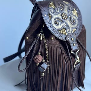 Pu&ograve; includere: Uno zaino in pelle marrone scuro con lunghe frange, una patta superiore con un ornato motivo dorato in rilievo e ciondoli decorativi in pelle intrecciata con piccole perline metalliche e dettagli cubici appesi sul lato.