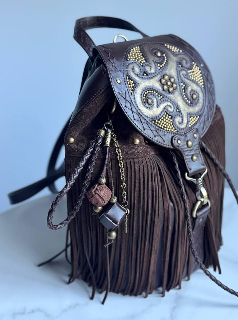 Pu&ograve; includere: Uno zaino in pelle marrone scuro con lunghe frange, una patta superiore con un ornato motivo dorato in rilievo e ciondoli decorativi in pelle intrecciata con piccole perline metalliche e dettagli cubici appesi sul lato.