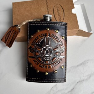 May include: A leather flask with a pirate design. The flask is black and brown, featuring a skull and crossbones image and the text "THINK LIKE A CAPTAIN ACT LIKE A PIRATE". It is presented in a brown box with a "hand made" tag.