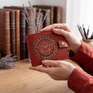 May include: A red leather wallet featuring a circular embossed design of a tree within an intricate Celtic knot border. The wallet includes a small tab closure secured with a brass-colored metal snap button on the right side of the front panel.