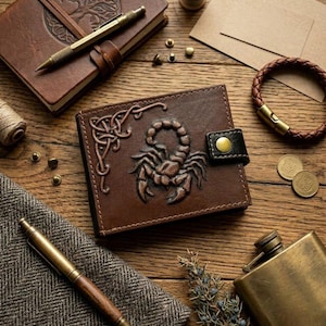 Pode incluir: Uma carteira de couro marrom com um design de escorpião é o ponto focal desta imagem. Outros itens incluem um diário encadernado em couro, uma caneta, uma pulseira, um frasco e moedas, todos dispostos em uma superfície de madeira.