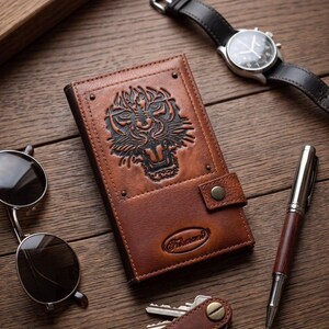 Puede incluir: Una cartera de cuero marrón con un diseño detallado de cara de tigre, un reloj negro, gafas de sol, un bolígrafo y llaves sobre una superficie de madera. La cartera tiene un cierre de botón a presión.