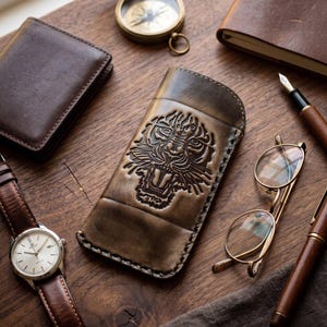 Puede incluir: Un estuche de cuero marrón para gafas con un diseño de cabeza de tigre en relieve en la parte frontal. El estuche está rodeado por una cartera de cuero marrón, una brújula de latón, una pluma estilográfica, un reloj de pulsera y gafas doradas.