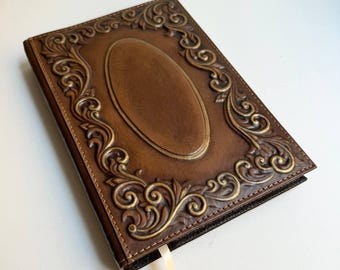 Pode incluir: Um diário de couro marrom com um design ornamentado e em relevo. A capa apresenta um grande oval no centro e uma borda decorativa.