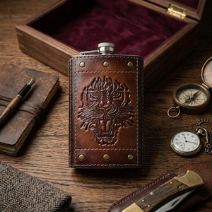 Puede incluir: Un petaca de cuero marrón con un diseño detallado de cara de tigre. La petaca tiene remaches de latón y una tapa plateada. Otros elementos en la superficie de madera incluyen una brújula, un reloj de bolsillo y un cuchillo pequeño.