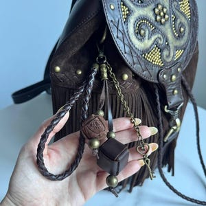 Pu&ograve; includere: Uno zaino in pelle marrone scuro con lunghe frange, una patta con un motivo dorato in rilievo e borchie in ottone. Un set di ciondoli include un cordino in pelle intrecciata, un piccolo cubo e un ciondolo a forma di chiave con cuore.