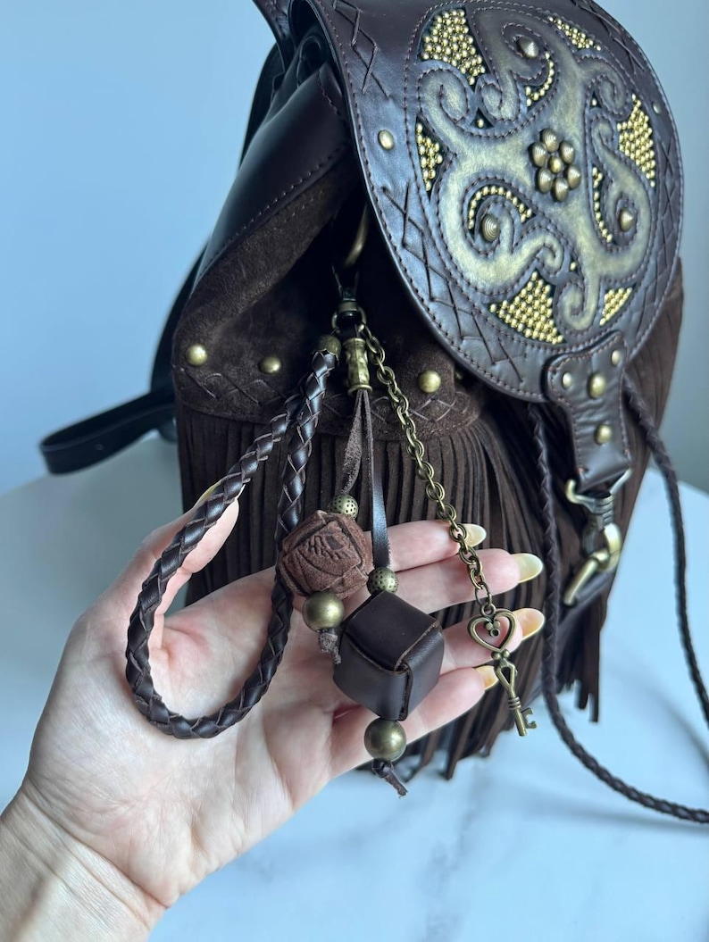 Pu&ograve; includere: Uno zaino in pelle marrone scuro con lunghe frange, una patta con un motivo dorato in rilievo e borchie in ottone. Un set di ciondoli include un cordino in pelle intrecciata, un piccolo cubo e un ciondolo a forma di chiave con cuore.