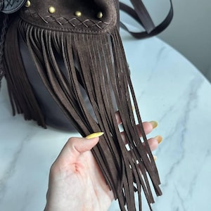 Pu&ograve; includere: Una borsa in camoscio marrone scuro con lunghe frange, una patta decorativa con cuciture complesse e piccoli borchie in metallo color ottone lungo il bordo superiore. La borsa include una tracolla sottile e una struttura in pelle liscia.