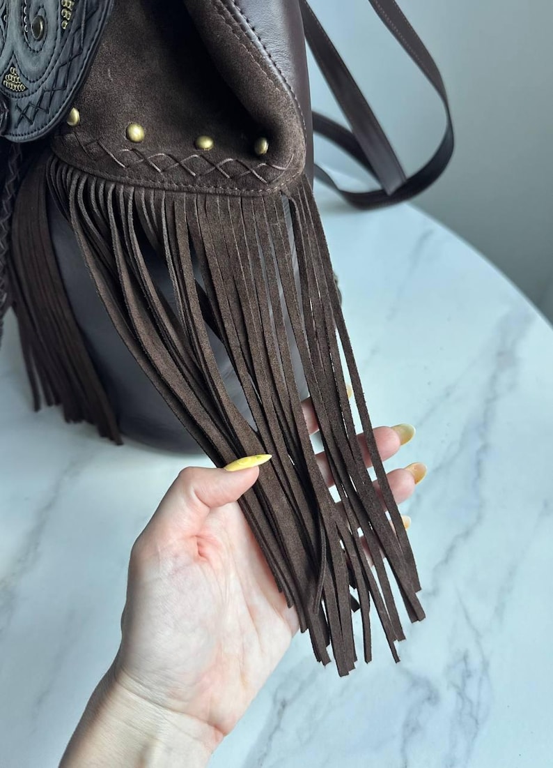 Pu&ograve; includere: Una borsa in camoscio marrone scuro con lunghe frange, una patta decorativa con cuciture complesse e piccoli borchie in metallo color ottone lungo il bordo superiore. La borsa include una tracolla sottile e una struttura in pelle liscia.