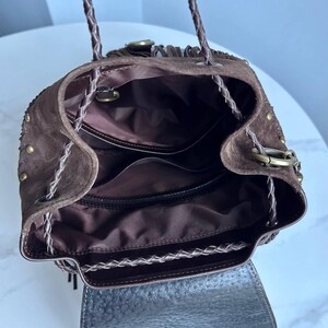 Pu&ograve; includere: Uno zaino in camoscio marrone scuro con chiusura a coulisse, spallacci in pelle intrecciata, borchie in metallo color ottone sui lati e un interno foderato in tessuto marrone scuro con scomparti. L'esterno presenta una patta e frange.