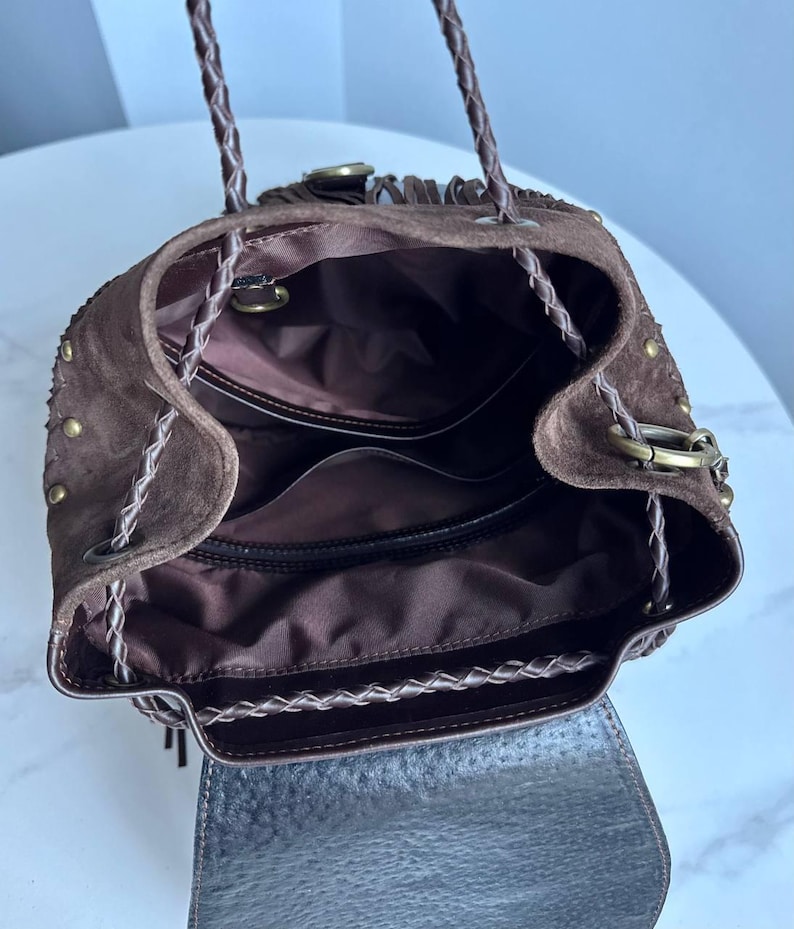 Pu&ograve; includere: Uno zaino in camoscio marrone scuro con chiusura a coulisse, spallacci in pelle intrecciata, borchie in metallo color ottone sui lati e un interno foderato in tessuto marrone scuro con scomparti. L'esterno presenta una patta e frange.