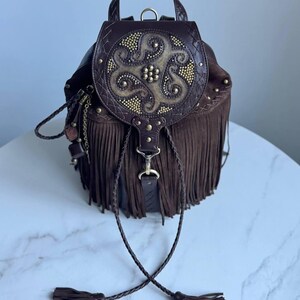 Pu&ograve; includere: Uno zaino in pelle marrone scuro con lunghe frange, un manico superiore e una patta circolare decorata con intricati rilievi e piccoli borchie metalliche dorate. La borsa include cordini in pelle intrecciata con nappe.