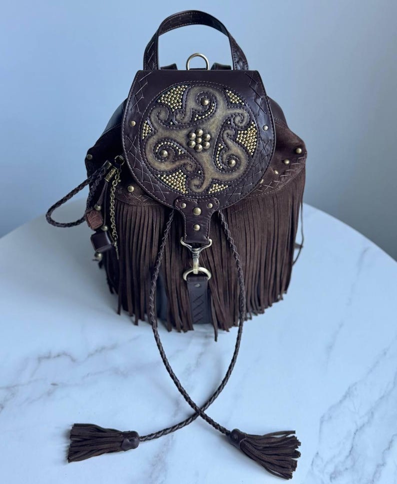 Pu&ograve; includere: Uno zaino in pelle marrone scuro con lunghe frange, un manico superiore e una patta circolare decorata con intricati rilievi e piccoli borchie metalliche dorate. La borsa include cordini in pelle intrecciata con nappe.