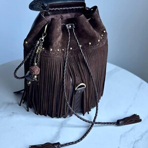 Pu&ograve; includere: Una borsa a secchiello in camoscio marrone scuro con lunghe frange, chiusura a coulisse intrecciata con nappe e borchie in metallo color ottone anticato. La borsa include un ciondolo decorativo con una piccola sfera e una patta in pelle scura.