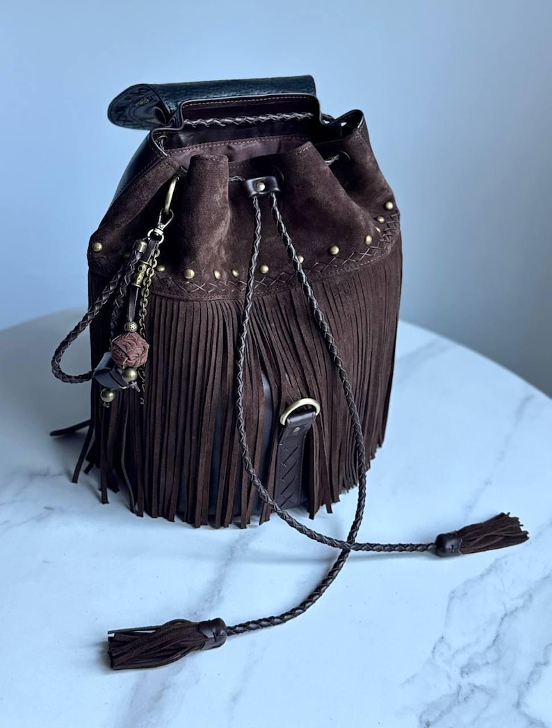 Pu&ograve; includere: Una borsa a secchiello in camoscio marrone scuro con lunghe frange, chiusura a coulisse intrecciata con nappe e borchie in metallo color ottone anticato. La borsa include un ciondolo decorativo con una piccola sfera e una patta in pelle scura.