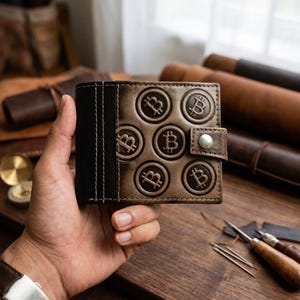 May include: A bi-fold leather wallet featuring a dark brown section and a lighter brown section embossed with multiple circular Bitcoin symbols. The wallet includes a small strap closure secured with a silver-toned metal snap button fastener.