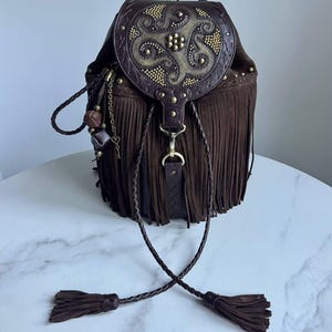 Pu&ograve; includere: Uno zaino in camoscio marrone scuro con lunghe frange, una patta superiore con un decorativo design circolare in rilievo e borchie metalliche. La borsa include cordini in pelle intrecciata con nappe e una catenella con ciondoli in metallo.
