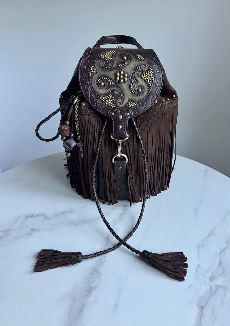 Pu&ograve; includere: Uno zaino in camoscio marrone scuro con lunghe frange, una patta superiore con un decorativo design circolare in rilievo e borchie metalliche. La borsa include cordini in pelle intrecciata con nappe e una catenella con ciondoli in metallo.