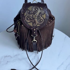 Pu&ograve; includere: Uno zaino in pelle marrone scuro con lunghe frange, un manico superiore e una patta circolare decorata con borchie metalliche dorate e un motivo centrale in rilievo. La borsa include lacci in pelle intrecciata con nappe.