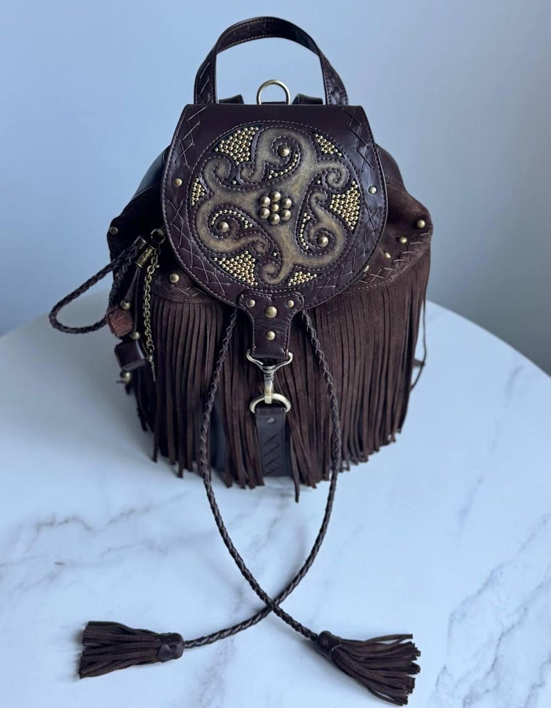 Pu&ograve; includere: Uno zaino in pelle marrone scuro con lunghe frange, un manico superiore e una patta circolare decorata con borchie metalliche dorate e un motivo centrale in rilievo. La borsa include lacci in pelle intrecciata con nappe.