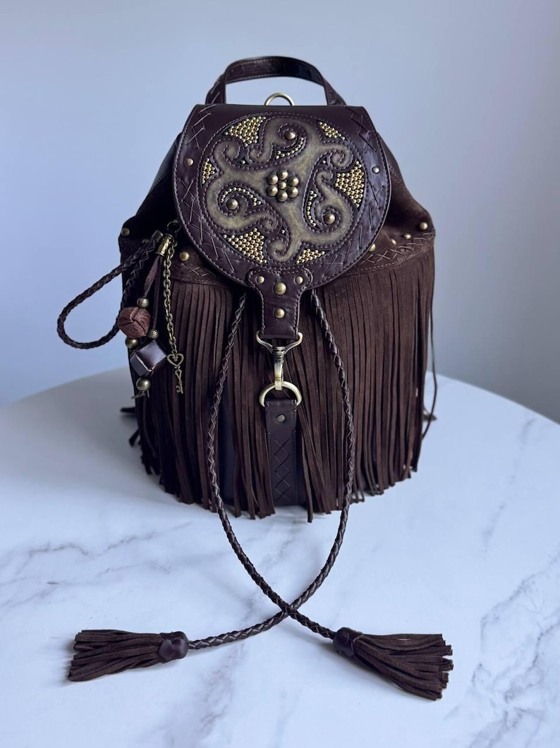 Pu&ograve; includere: Uno zaino in camoscio marrone scuro con lunghe frange, un manico superiore e una patta circolare decorata con borchie metalliche dorate e motivi in rilievo. La borsa include una chiusura a coulisse intrecciata con nappe e ciondoli in metallo.