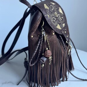 Pu&ograve; includere: Uno zaino in pelle marrone scuro con lunghe frange, chiusura a patta con borchie dorate e un set di ciondoli decorativi composto da cordini in pelle intrecciata, catene metalliche, perline e un piccolo pendente a forma di chiave.