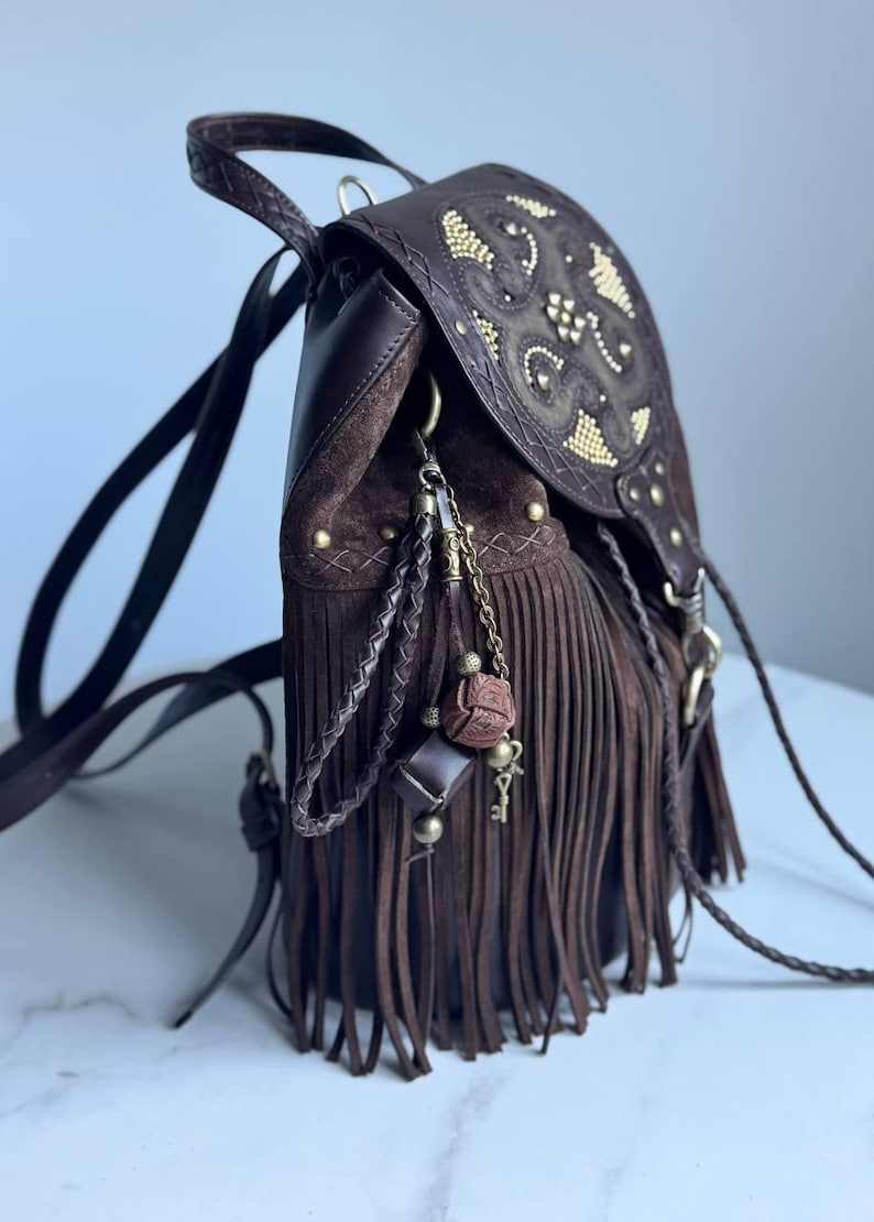 Pu&ograve; includere: Uno zaino in pelle marrone scuro con lunghe frange, chiusura a patta con borchie dorate e un set di ciondoli decorativi composto da cordini in pelle intrecciata, catene metalliche, perline e un piccolo pendente a forma di chiave.