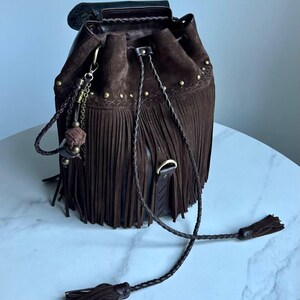 Pu&ograve; includere: Una borsa a secchiello in camoscio marrone scuro con lunghe frange, chiusura a coulisse intrecciata con nappe e minuteria metallica color ottone antico. La borsa presenta un ciondolo decorativo e un bordo in pelle intrecciata sulla parte superiore.