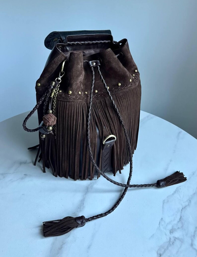 Pu&ograve; includere: Una borsa a secchiello in camoscio marrone scuro con lunghe frange, chiusura a coulisse intrecciata con nappe e minuteria metallica color ottone antico. La borsa presenta un ciondolo decorativo e un bordo in pelle intrecciata sulla parte superiore.