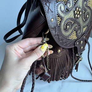 Pu&ograve; includere: Uno zaino in pelle marrone scuro con una patta decorata da un motivo dorato in rilievo e piccole borchie dorate. La borsa presenta lunghe frange in camoscio, cinturini in pelle intrecciata e un gancio a moschettone in metallo con catenella.