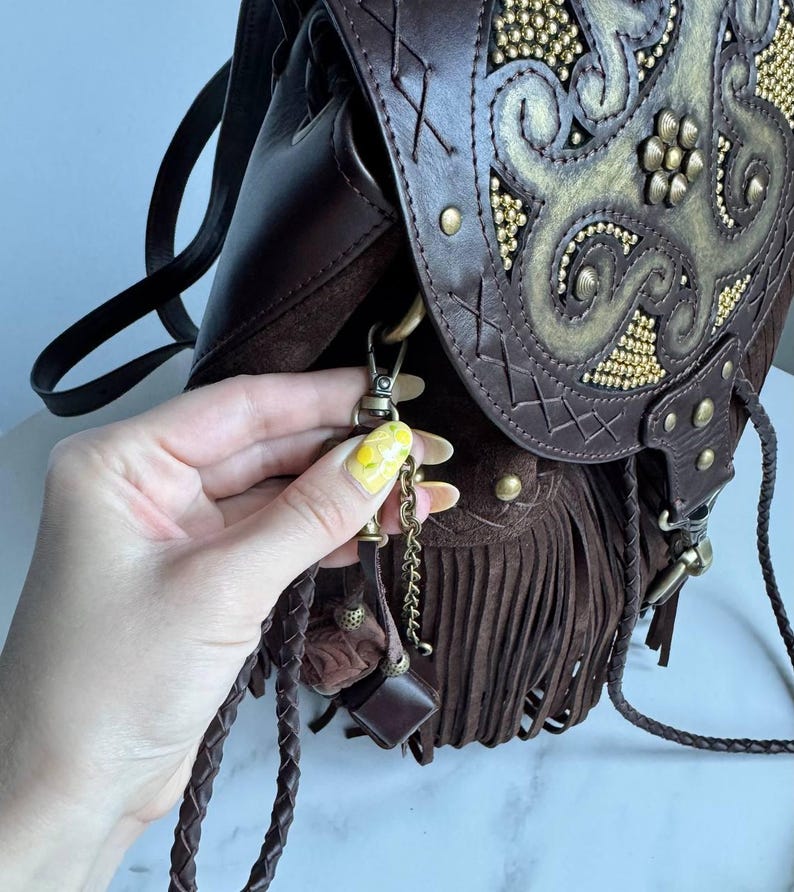 Pu&ograve; includere: Uno zaino in pelle marrone scuro con una patta decorata da un motivo dorato in rilievo e piccole borchie dorate. La borsa presenta lunghe frange in camoscio, cinturini in pelle intrecciata e un gancio a moschettone in metallo con catenella.