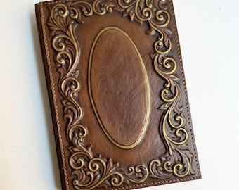 Pode incluir: Um diário de couro marrom com um design em relevo dourado. O diário tem uma moldura oval decorativa com detalhes florais ornamentados.