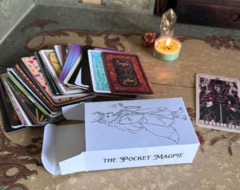 Könnte beinhalten: Eine offene weiße Schachtel mit dem Text "THE POCKET MAGPIE" neben einem Tarotkartenspiel. Die Karten haben verschiedene Designs und Farben. Eine brennende Kerze und ein Kristall sind im Hintergrund.