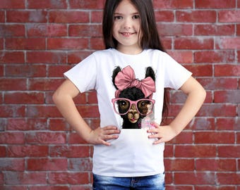May include: White t-shirt featuring a graphic of a black llama wearing a pink bow, glasses, and a pink scarf. The llama is centered on the shirt. The girl is wearing blue jeans and is standing in front of a red brick wall.