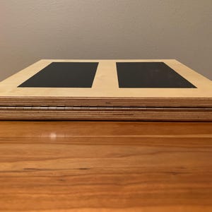 May include: A wooden laptop desk with a natural finish and two black rectangular pads. The desk is folded, revealing a metal hinge. The desk is designed to be portable and adjustable for comfortable use.